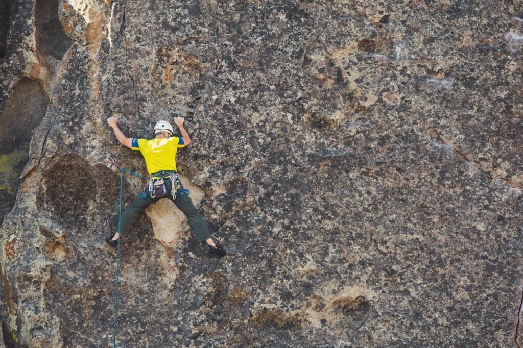 A climber in a helmet and safety gear scales a rough rock face in daylight.